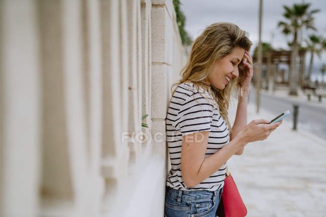 Вид сбоку счастливой женщины в повседневной полосатой рубашке и джинсах, стоящей рядом со зданием на городской улице и использующей смартфон — стоковое фото