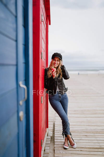 Trendfrau in schwarzer Mütze und Lederjacke isst einen roten Apfel, während sie sich an eine hölzerne Strandkabine lehnt — Stockfoto