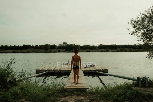 Vista posteriore del bambino sportivo attivo che si sposta sul molo verso l'acqua pacifica del lago durante la giornata estiva — Foto stock