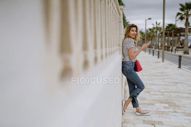 Вид збоку жінки, що стоїть поруч з будівлею на міській вулиці, використовуючи смартфон і дивлячись на камеру — стокове фото