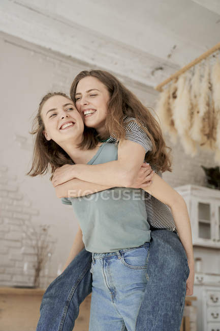 Cheerful casual women laughing and having fun at home as riding piggy back — Stock Photo