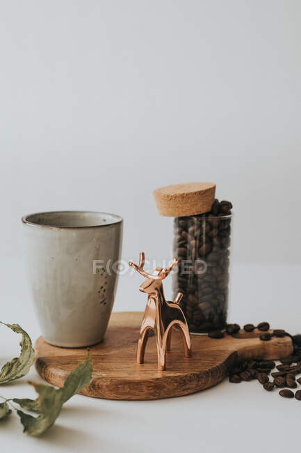 Figurinha de veado brilhante composta com jarra de grãos de café e caneca em suporte de madeira em fundo cinza — Fotografia de Stock