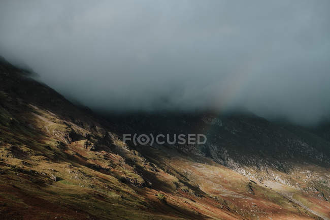 Paisaje salvaje de colinas rocosas cubiertas de espesa niebla y tenue arco iris en Escocia - foto de stock