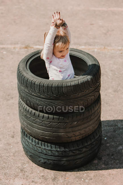 Зверху щаслива чарівна дівчинка стоїть в стосі автомобільних шин, розважаючись і граючи на відкритому повітрі в літній день — стокове фото
