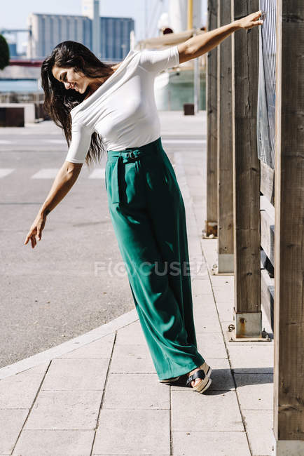 Hermosa joven sonriente colgando en un poste en la calle mientras mira hacia abajo - foto de stock