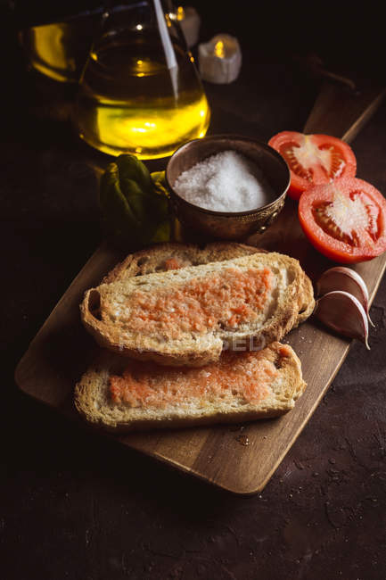 Various spices and ripe tomatoes placed on cutting board near pieces of bread with sauce on table — Stock Photo