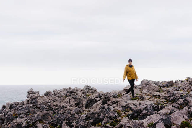 Жінка в жовтому капюшоні ступає на кам'яний берег, оточений пінявими хвилями в сірий хмарний день. — Stock Photo