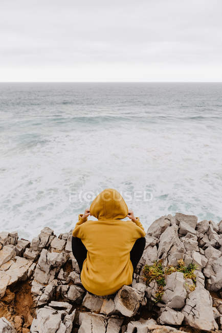 Vista trasera del viajero con capucha amarilla cálida sentado solo en la orilla rocosa mirando las olas espumosas en el día nublado - foto de stock