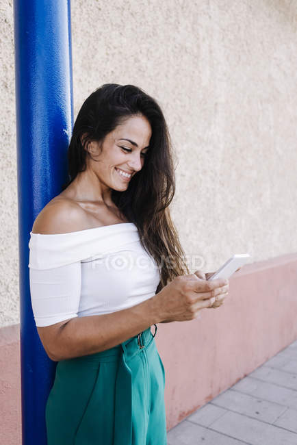 Счастливая молодая женщина с длинными волосами, прислонившаяся к стене, смотрит на мобильный телефон — стоковое фото