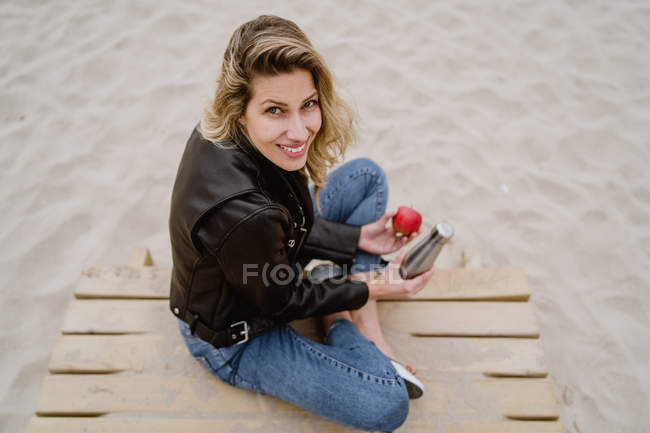 De cima mulher loira na moda em jaqueta de couro comendo maçã madura vermelha na praia de areia olhando para a câmera — Fotografia de Stock