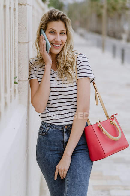 Mujer alegre feliz en camisa de rayas casuales y jeans de pie junto al edificio en la calle de la ciudad y hablando en el teléfono inteligente - foto de stock