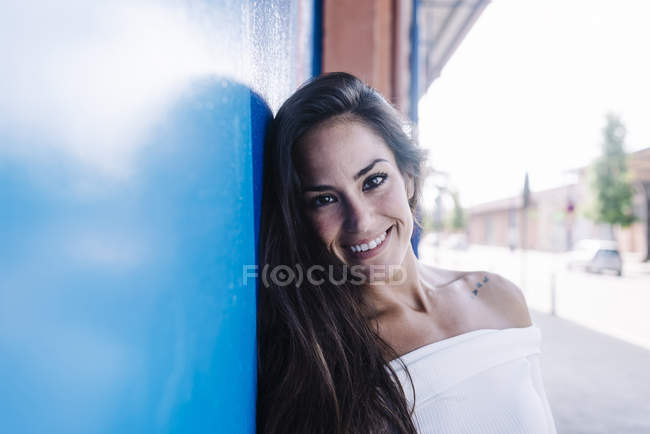 Giovane donna sorridente appoggiata a un muro blu guardando la fotocamera — Foto stock