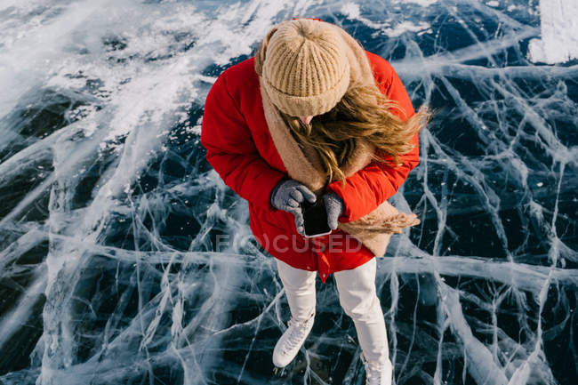 From above of modern active female in warm clothes and skates standing on dark cracked ice on frozen river and using smartphone — Stock Photo