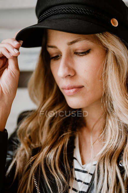 Stylische schöne selbstbewusste Frau in Lederjacke schaut weg, während sie die trendige schwarze Mütze im Kopf an der Holzwand berührt — Stockfoto