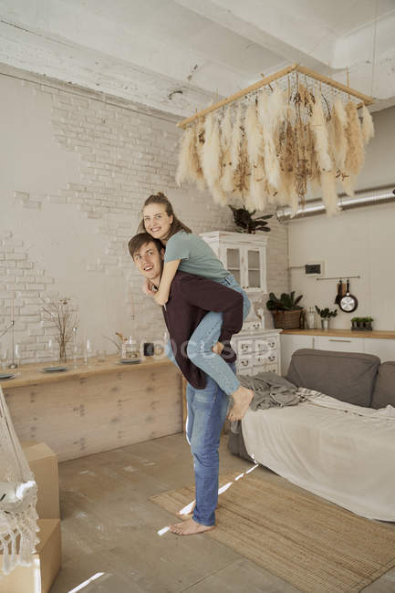 Vista lateral do homem bonito sorrindo dando passeio de piggyback para a fêmea alegre na sala de estar acolhedora luz — Fotografia de Stock