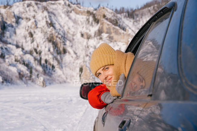 Счастливая улыбающаяся молодая женщина, сидящая в машине и наклоняющаяся в окно во время дорожной поездки в заснеженные горы в Сибири, Россия — стоковое фото