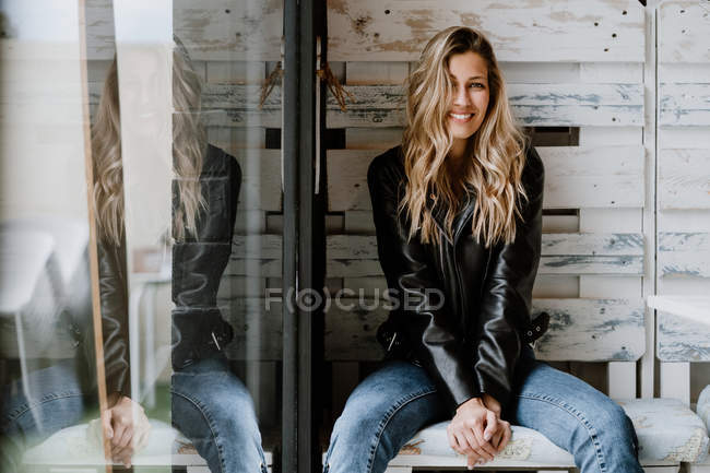 Trendy langhaarige Frau in Lederjacke und Mütze sitzt auf einer Holzbank und blickt in die Kamera — Stockfoto