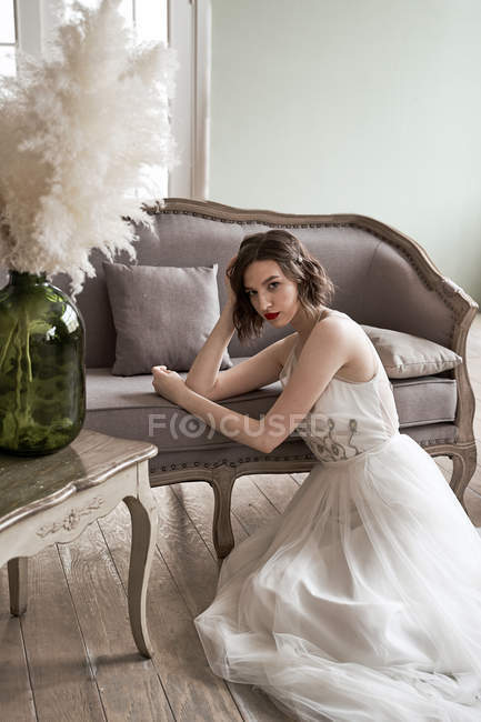 Mulher linda com lábios vermelhos em vestido branco olhando para a câmera enquanto sentado no chão ao lado do sofá — Fotografia de Stock