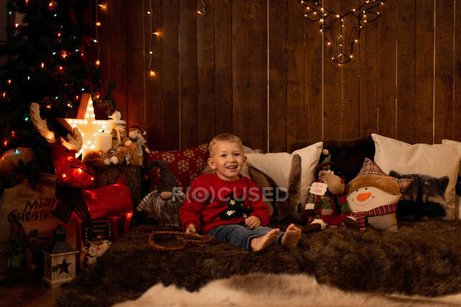 Adorable petit garçon assis dans la chambre pleine de décoration de Noël et regardant à la caméra — Photo de stock