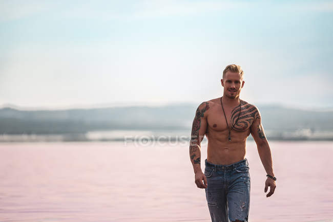 Muscular tattooed shirtless guy in jeans strolling around pink lake at Torrevieja on blurred background — Stock Photo