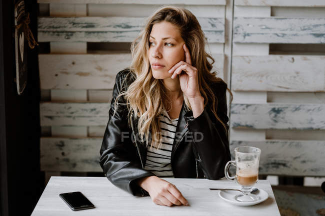 Stylish long haired blonde in black leather jacket looking away at table in cafe — Stock Photo