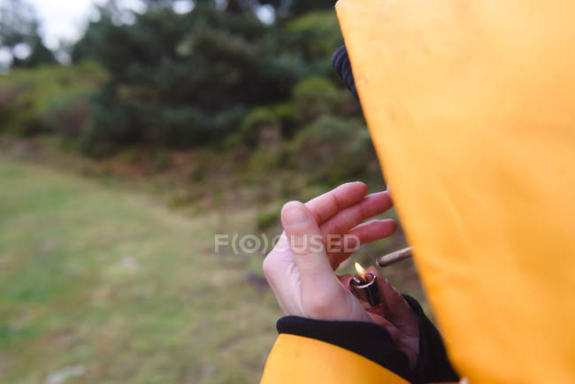 Обрезанное изображение женщины в желтом дождевике, освещающей сигарету в лесу — стоковое фото