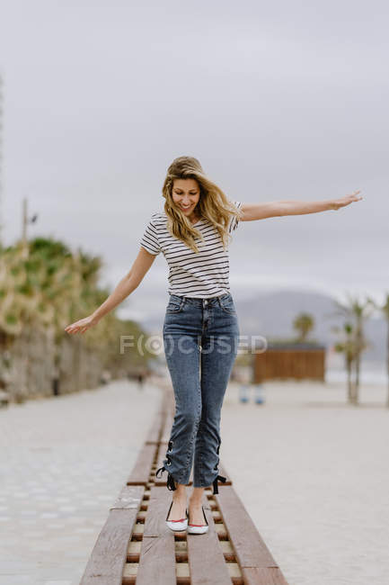 Happy cheerful woman in casual clothes walking with arms spread on wooden bench and enjoying summer day at city seafront — Stock Photo