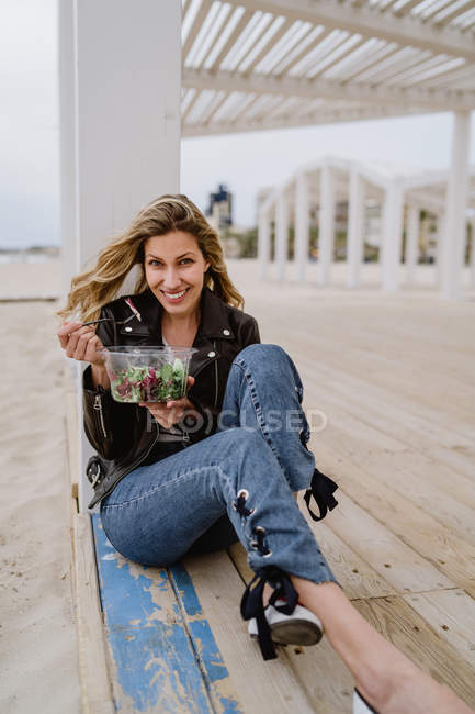 Stylish woman in black jacket enjoying healthy green salad while sitting on wooden terrace on coast smiling at camera — Stock Photo