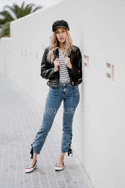 Trendy long haired blonde in black cap and jacket with denim confidently looking at camera and leaning on wall — Stock Photo