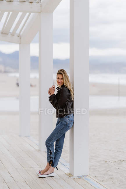 Side view of woman in black leather jacket confidently looking at camera and leaning on white pillar in cloudy day — Stock Photo