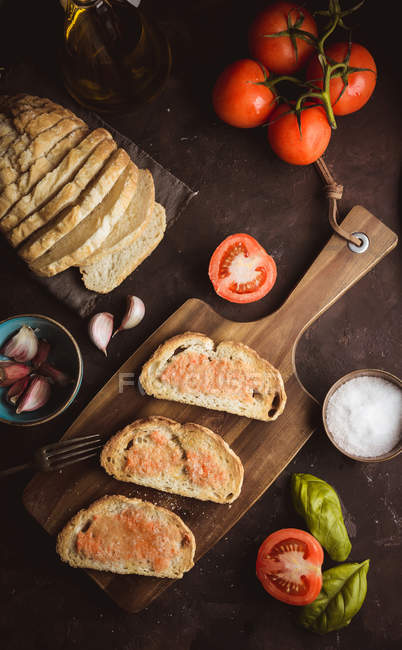 Diverses épices et tomates mûres placées sur une planche à découper près de morceaux de pain avec sauce sur la table — Photo de stock