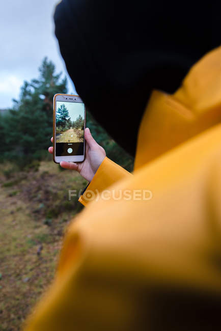 Frau in Kapuzenpulli und gelbem Regenmantel fotografiert Wald mit Smartphone — Stockfoto