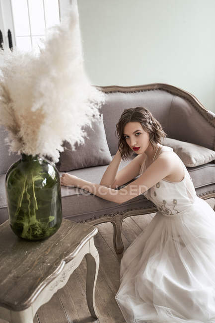 Зверху чудова спокійна жінка в елегантній білій весільній сукні, що торкається волосся і дивиться на камеру, сидячи на підлозі біля елегантного дивана в старовинному стилі — стокове фото