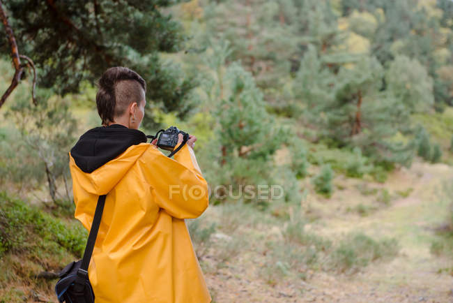 Back view of woman in yellow raincoat photographing tree in mountain — Stock Photo