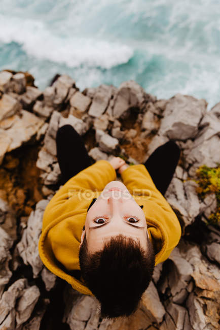 Von oben eine Frau in gelbem warmen Kapuzenpulli, die allein am felsigen Ufer mit schäumenden Wellen an bewölkten Tagen sitzt und in die Kamera blickt — Stockfoto