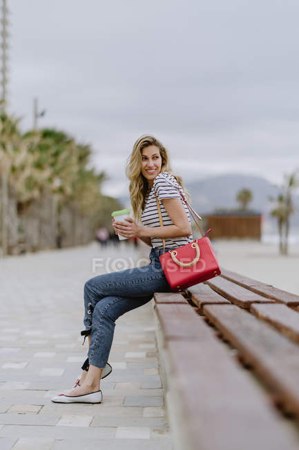 Belle femme moderne avec un beau sourire portant une chemise rayée décontractée regardant loin tout en étant assis sur le banc — Photo de stock