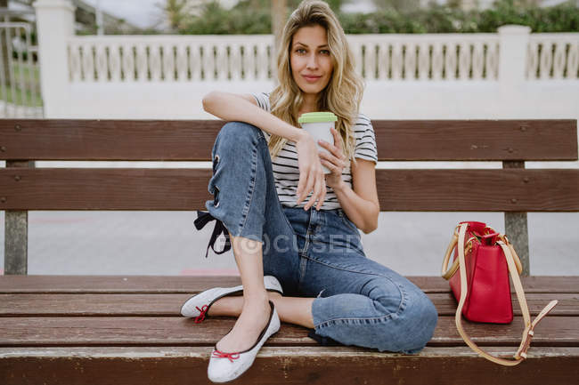 Femme avec tasse à emporter de café assis sur le banc de la ville en bord de mer le jour de l'été — Photo de stock