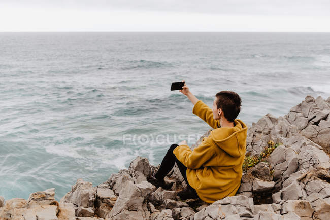 Donna dai capelli corti in felpa gialla seduta sulla spiaggia rocciosa e scattare selfie sul telefono cellulare nella giornata nuvolosa grigia — Foto stock