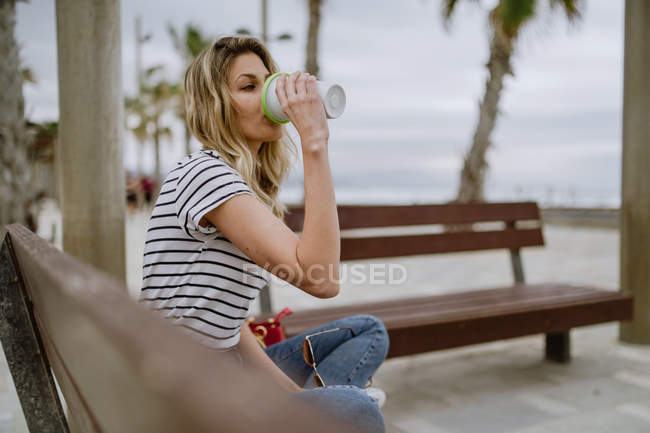 Вид сбоку женщины, пьющей из чашки кофе, сидящей на городской скамейке у моря в летний день — стоковое фото