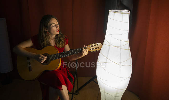 Зверху талановита жінка в червоній сукні виконує пісню і грає на гітарі в теплій освітленій сцені поблизу білої лампи — стокове фото