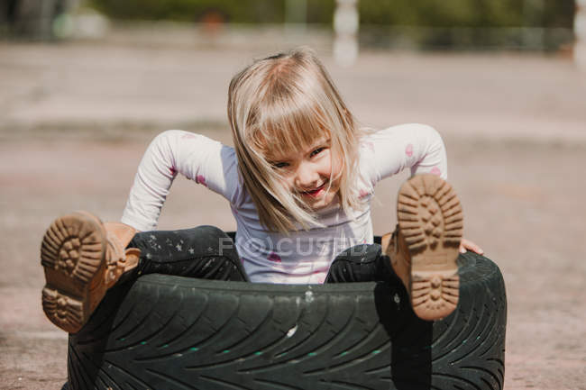 Зверху щаслива чарівна дівчинка сидить всередині автомобільної шини, розважаючись і граючи на відкритому повітрі в літній день — стокове фото
