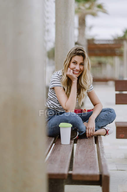 Cheerful casual young woman sitting on city bench at seafront on summer day looking at camera — Stock Photo