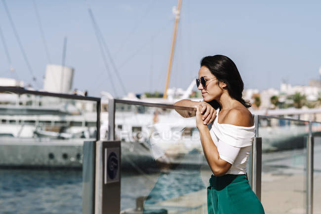 Seitenansicht einer verträumten jungen Frau mit Sonnenbrille, die am Zaun lehnt und am Hafendock wegschaut — Stockfoto