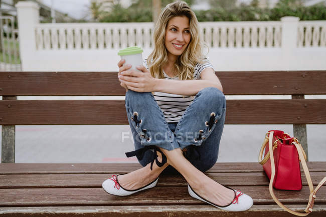 Femme occasionnelle joyeuse avec tasse de café à emporter assis sur le banc de la ville en bord de mer le jour de l'été — Photo de stock
