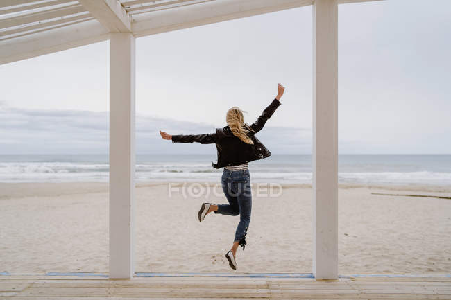 Back view of trendy woman in black jacket merrily jumping with arms raised on white wooden pier with ocean on background — Stock Photo