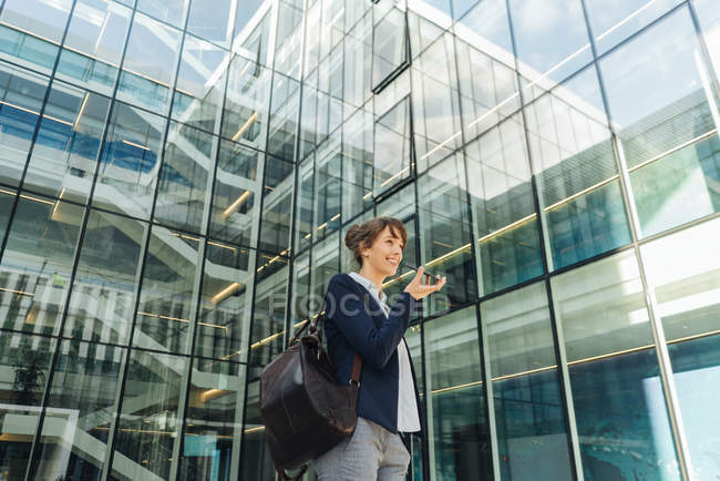 Низький кут щасливої бізнес-леді тримає руку в кишені і використовує смартфон, стоячи за межами бізнес-центру на міській вулиці — стокове фото
