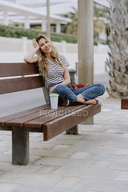 Vista lateral de la mujer alegre sentada en el banco de la ciudad en el paseo marítimo en el día de verano mirando a la cámara - foto de stock
