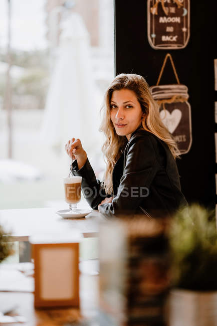 Side view of long haired trendy beautiful blonde woman drinking from a glass of delicious foamy coffee while looking at camera — Stock Photo