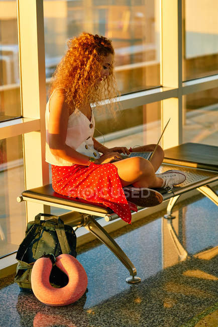 Elegante mujer alegre escribiendo en el ordenador portátil mientras está sentado con las piernas cruzadas en el banco de metal en el pasillo de cristal del aeropuerto de Texas - foto de stock