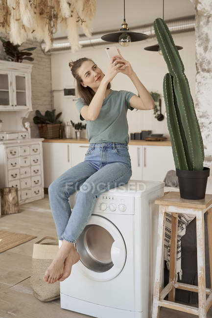 Chilling young barefoot woman having break on white and taking photo with mobile phone of cactus in kitchen — Stock Photo